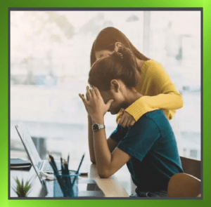 Person in distress at desk receiving support from a colleague who has placed a reassuring hand on their shoulder.
